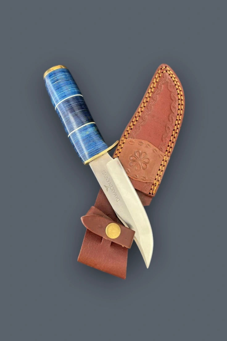 Diagonal flat lay of the Blazefury Hunter stainless steel hunting knife featuring a blue wood handle, resting across its cowhide leather sheath.