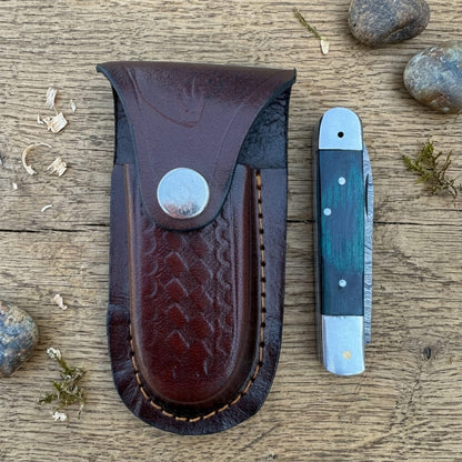 A dark brown leather knife sheath with a snap closure and stitched edge, displayed next to a folding pocket knife on a rustic wooden surface with small stones and wood shavings.