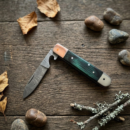 A folding pocket knife with a Damascus steel blade, a green inlay handle, and a copper-colored bolster, displayed on a rustic wooden surface with scattered stones and dried leaves.