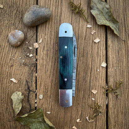 A folding pocket knife with a dark green and black marbled inlay handle, laid flat on a rustic wooden surface surrounded by small stones, dried leaves, and wood shavings.