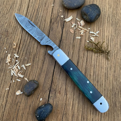 A folding pocket knife with a Damascus steel blade and a green inlay handle, displayed diagonally on a rustic wooden surface with small stones and wood shavings.