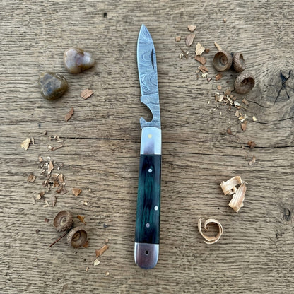A pocket knife with a patterned steel blade and a dark green to blue marbled handle, displayed on a rustic wooden surface with wood shavings and acorns.