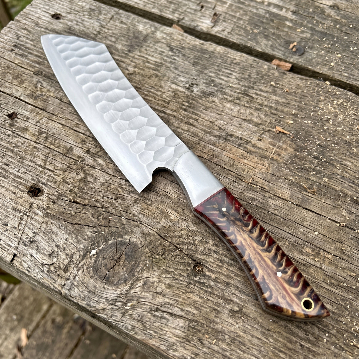 A custom chef's knife with a hammered Damascus steel blade and a decorative reddish-brown wood handle, resting diagonally on a rustic weathered wooden plank surface.