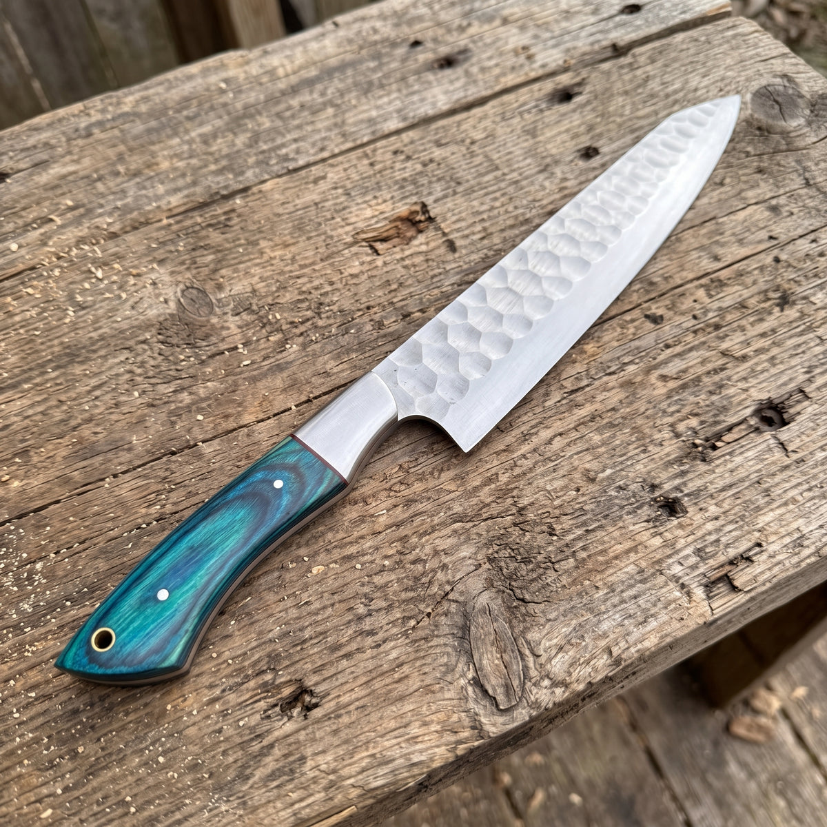 A chef's knife with a hammered Damascus steel blade and a marbled turquoise and green wooden handle, resting diagonally on a rustic weathered wooden surface.