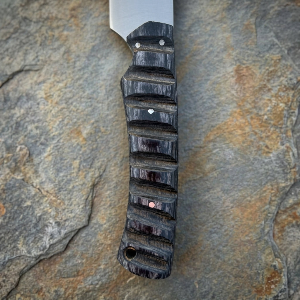 A close-up of a knife handle with a textured black and dark brown wood grain finish, featuring a silver blade and small metal pins, resting on a textured gray stone surface.