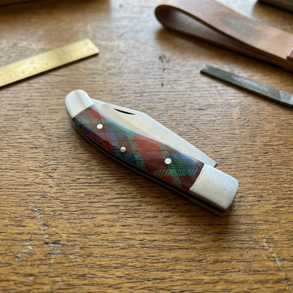 A old southern stainless steel pocket knife with a red, green, and blue tartan plaid handle, resting on a wooden workbench next to leather and metal tools.
