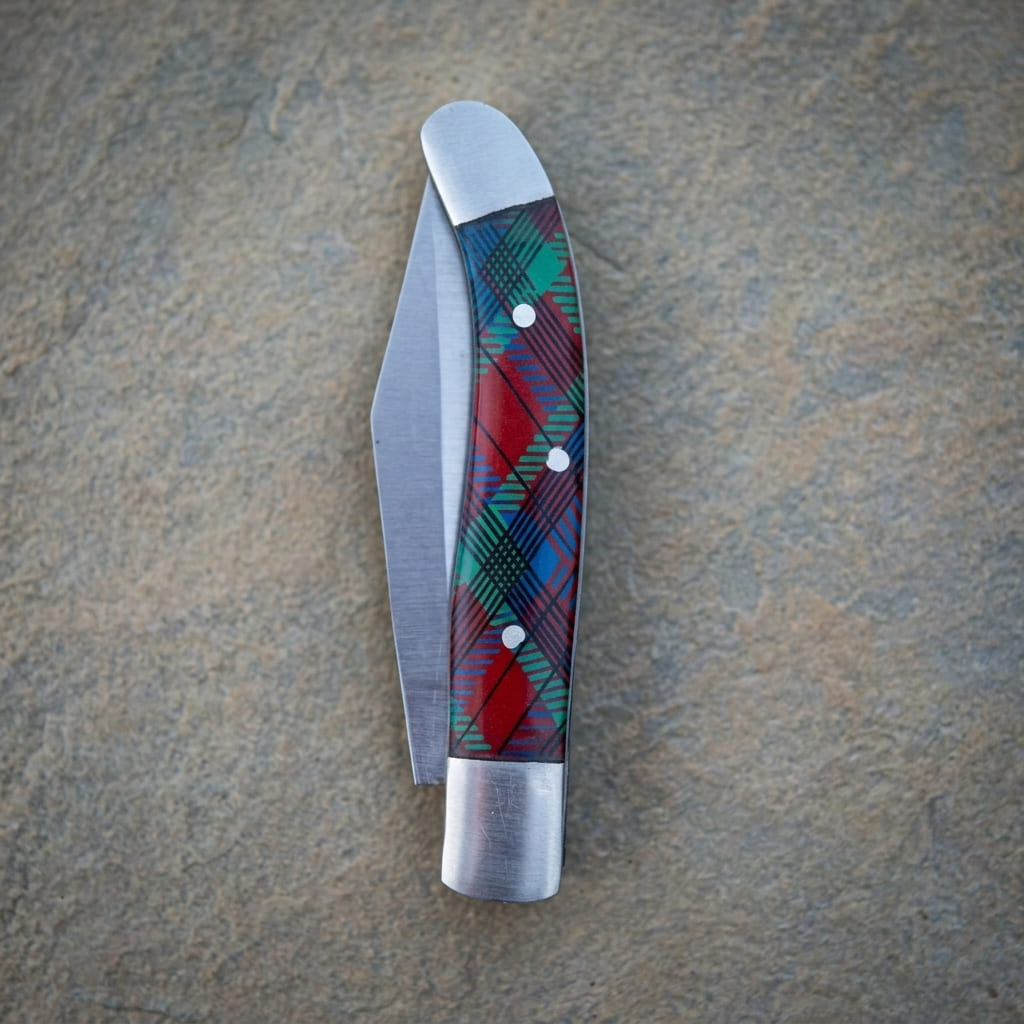 A classic old southern stainless steel pocket knife with a red, green, and blue tartan plaid handle, shown open on a textured gray stone surface.