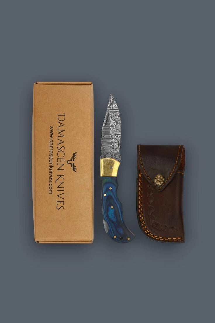 Vertical flat lay of the closed The Vanguard Damascus knife with blue pakkawood handle and brass bolsters.