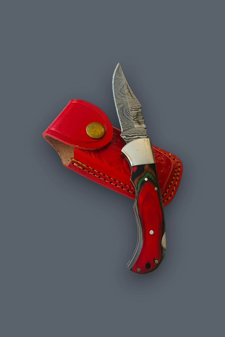 Fully open Olde Townie Damascus folding knife with a multicolor wood handle resting across its red leather sheath