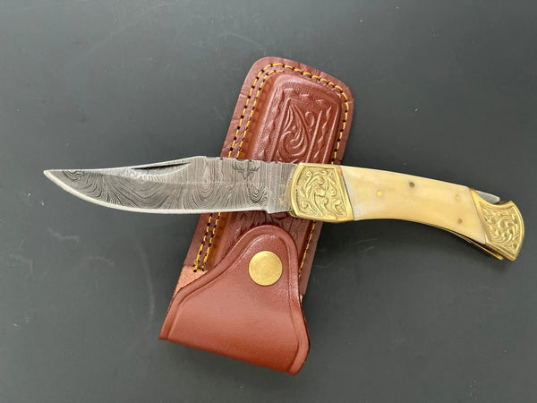 Damascus folding pocket knife with bone handle and 5-inch blade resting on premium leather sheath.