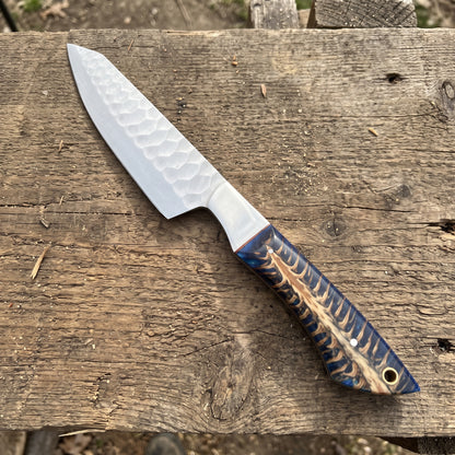 A fixed-blade knife with a hammered silver blade and a blue, brown, and gold patterned handle, resting diagonally on a rustic wooden plank.