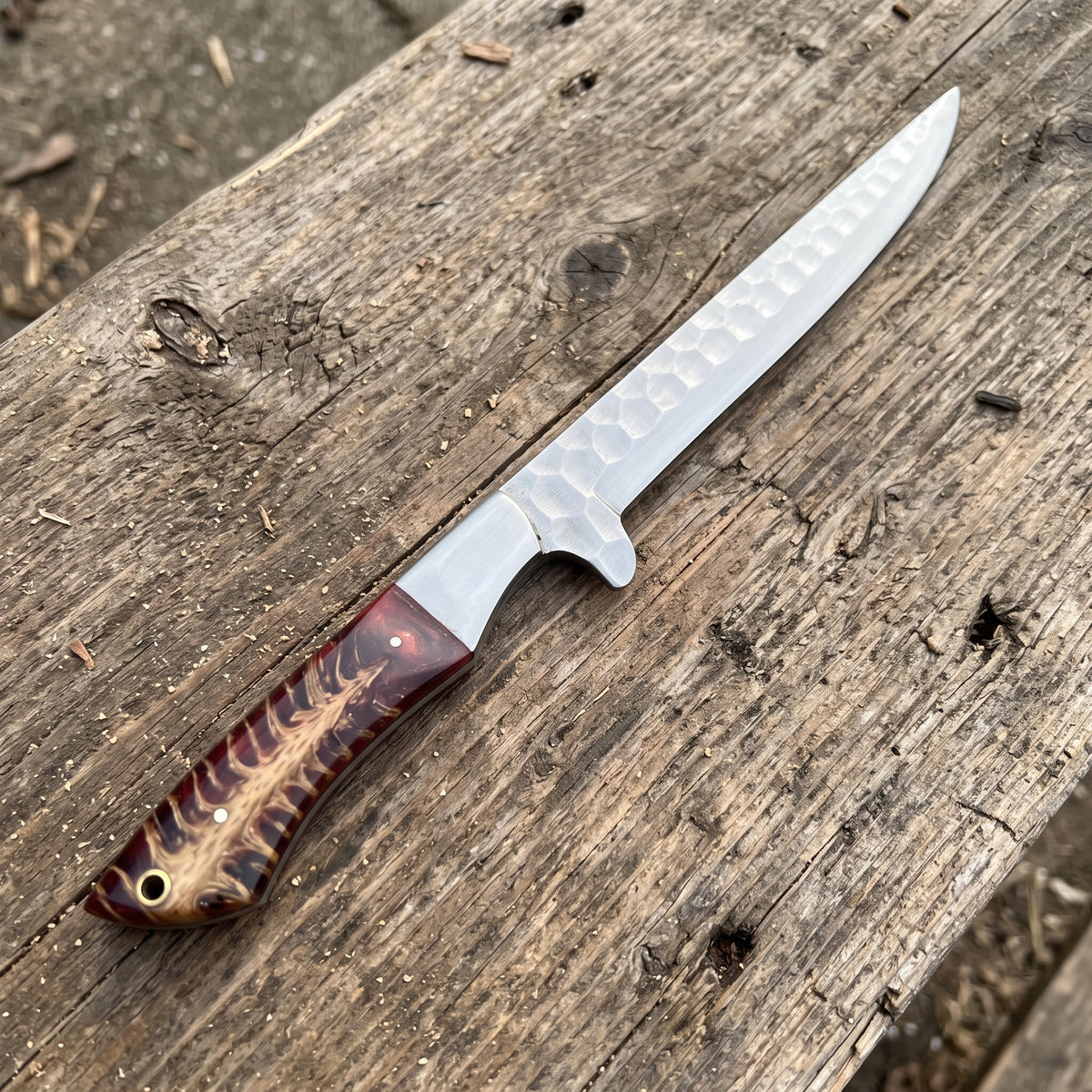 A fixed-blade knife with a hammered silver blade and a brown patterned wooden handle, resting diagonally on a weathered wooden plank outdoors.