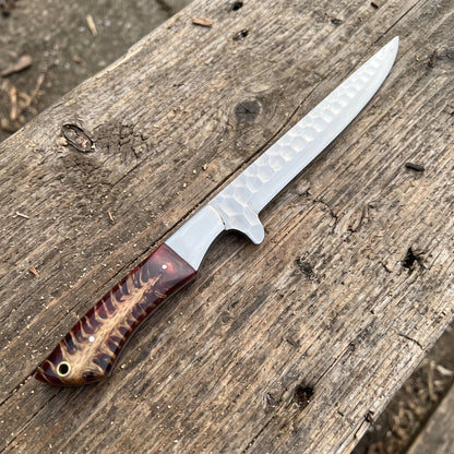 A fixed-blade knife with a hammered silver blade and a brown patterned wooden handle, resting diagonally on a weathered wooden plank outdoors.