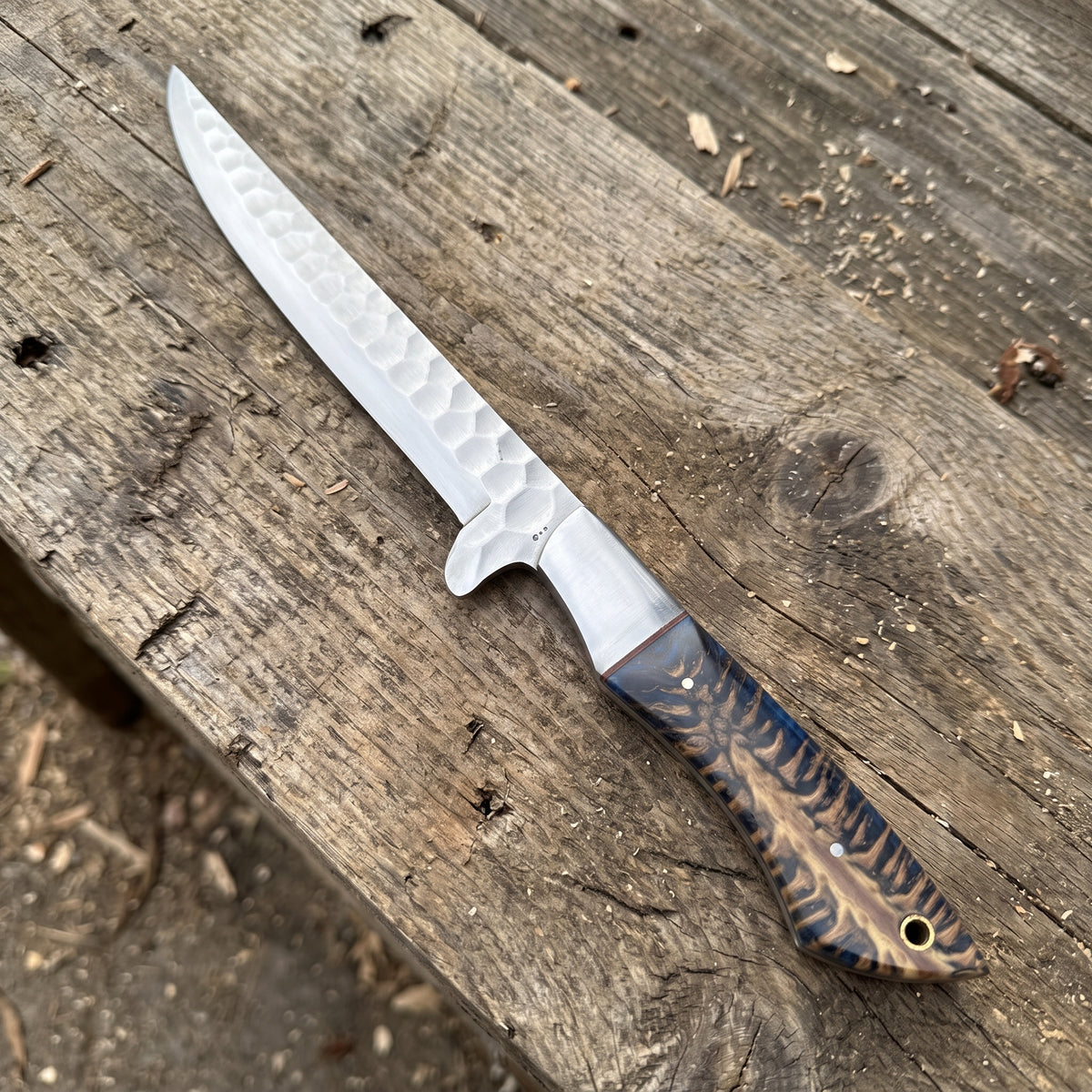 A fixed-blade knife with a hammered silver blade and a patterned brown and black handle, resting diagonally on a rustic weathered wooden surface.