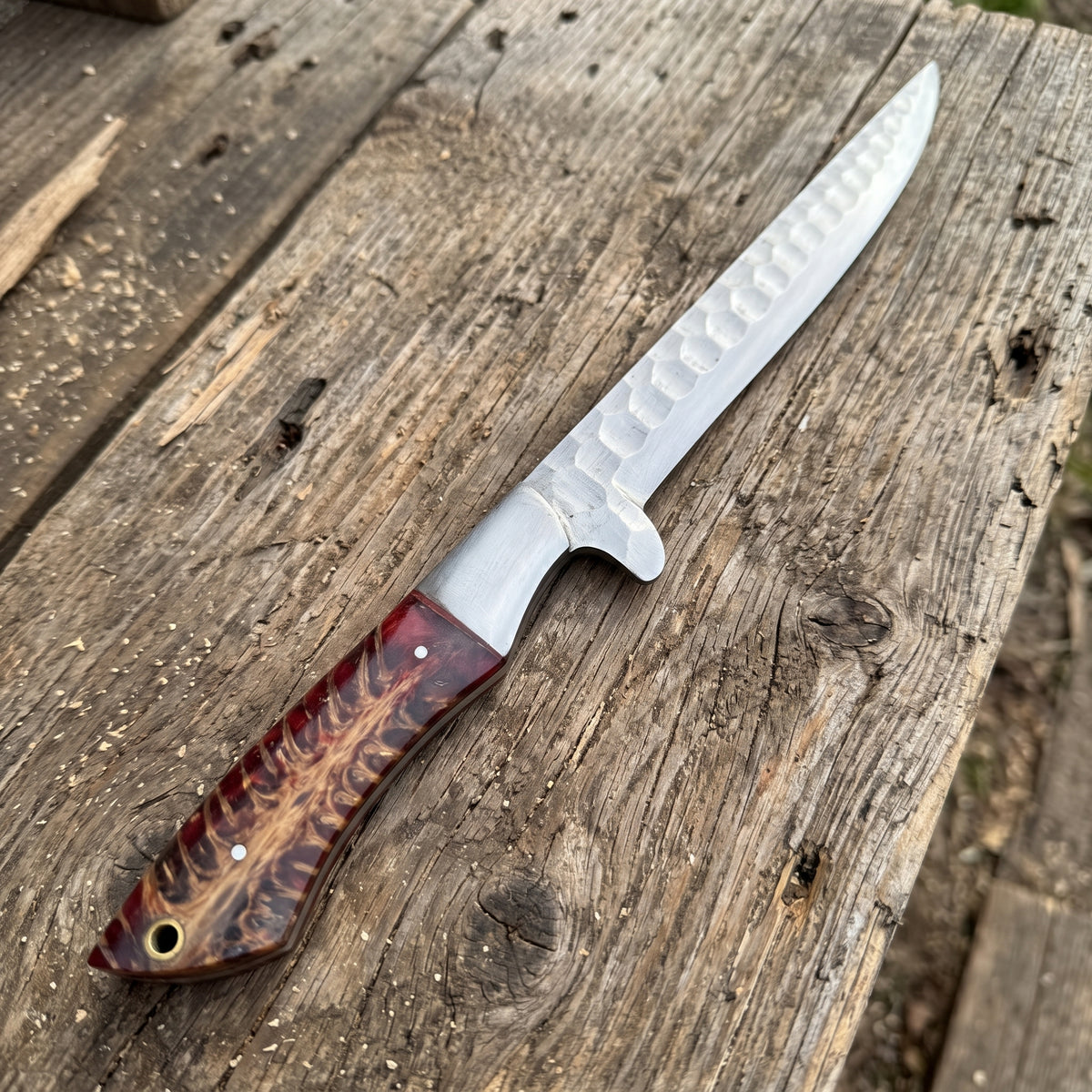 A fixed-blade knife with a hammered silver blade and a reddish-brown wood handle, resting diagonally on a rustic weathered wooden plank.