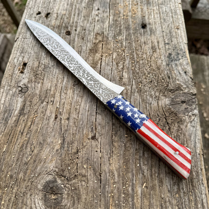 A fixed-blade knife with a patterned Damascus steel blade and an American flag handle, resting diagonally on a rustic wooden plank.