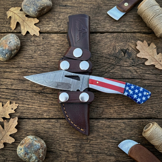 A fixed-blade knife with a patterned steel blade, silver hardware, and a handle featuring an American flag design, displayed in a dark brown leather sheath on a rustic wooden surface with autumn leaves and twine.