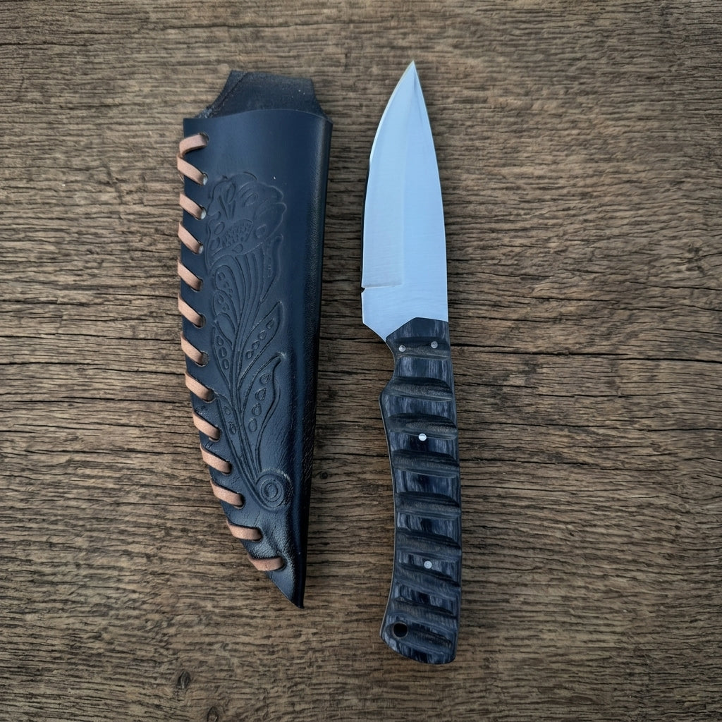 A fixed-blade knife with a silver blade and textured black handle, displayed next to its custom black leather sheath with tan stitching, set on a rustic wooden background.