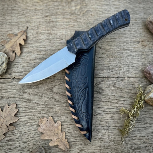 A fixed-blade knife with a silver steel blade and a textured black handle, shown partially sheathed in a black leather sheath with tan stitching, placed on a rustic wooden surface with scattered oak leaves.