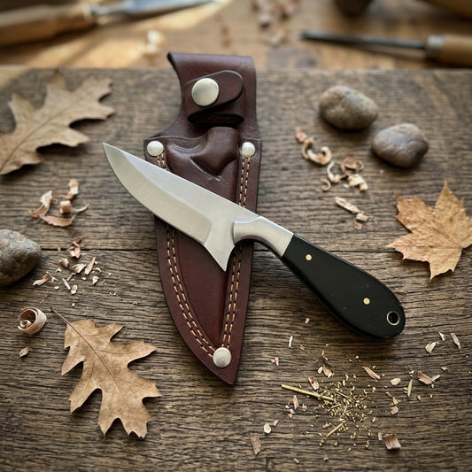 A fixed-blade knife with a silver steel blade and black handle lies on a brown leather sheath over a rustic wooden surface, surrounded by dried leaves, wood shavings, and small stones.