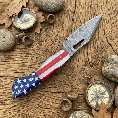 A fixed blade knife with a patterned steel blade and an American flag handle, displayed on a rustic wooden surface surrounded by stones, acorns, and vintage compasses.