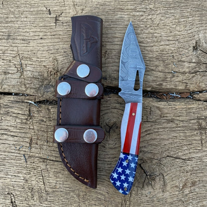 A custom fixed blade knife with a patterned steel blade and an American flag handle, displayed next to its brown leather sheath on a rustic wooden surface.