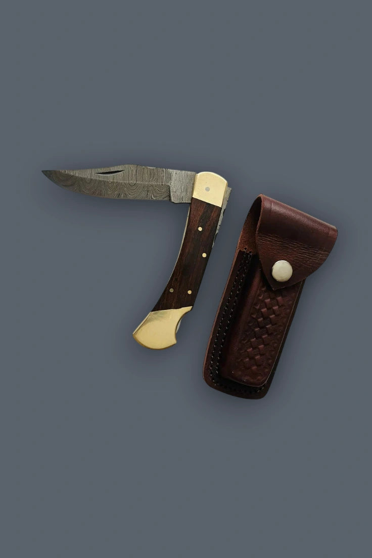 Diagonal flat lay of the folded Old World Edge Damascus knife featuring brass bolsters next to its stamped brown leather sheath.