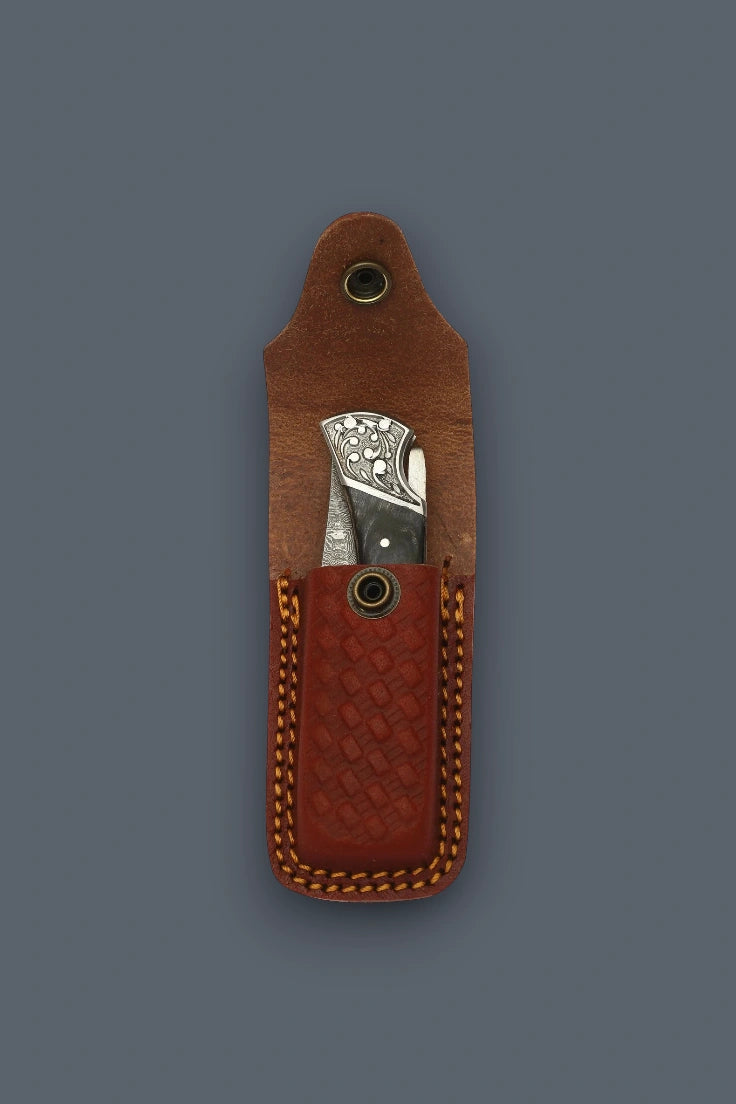 Folded The Squire Damascus pocket knife displaying its engraved silver bolster inside an open brown basketweave leather sheath.