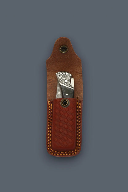 Folded The Squire Damascus pocket knife displaying its engraved silver bolster inside an open brown basketweave leather sheath.