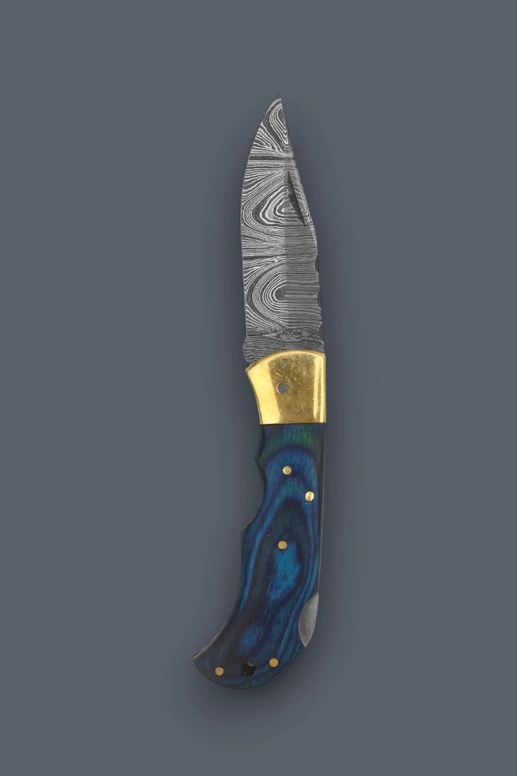 Diagonal flat lay of the folded The Vanguard Damascus knife featuring brass bolsters next to its stamped brown leather sheath.