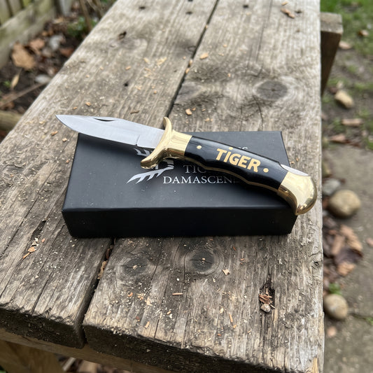 A folding pocket knife with a black and gold handle and a silver blade, resting on top of its black presentation box. The box features the brand name 'TIGER DAMASCEN'.