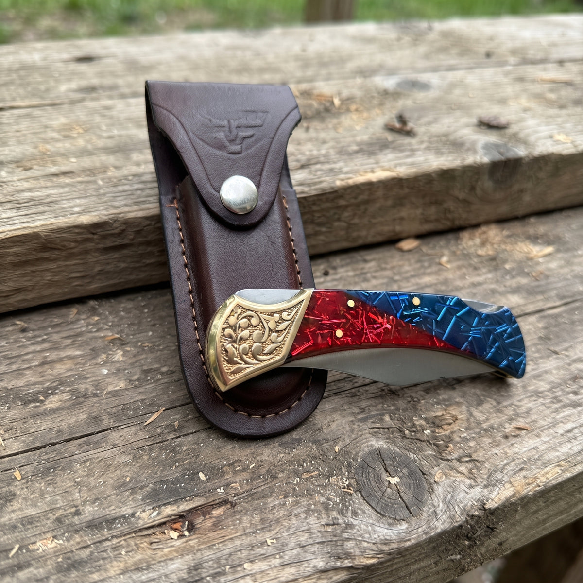 A folding pocket knife with a red and blue marbled handle and gold-toned guard, shown resting in a brown leather sheath on a rustic wooden surface.