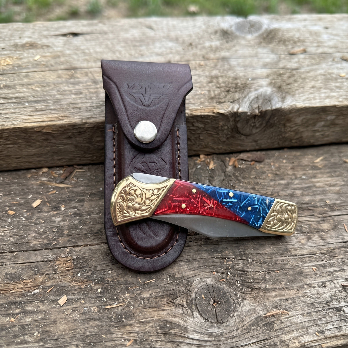 A folding pocket knife with a red and blue marbled handle and gold-toned hardware, resting in front of a brown leather sheath on a rustic wooden surface.