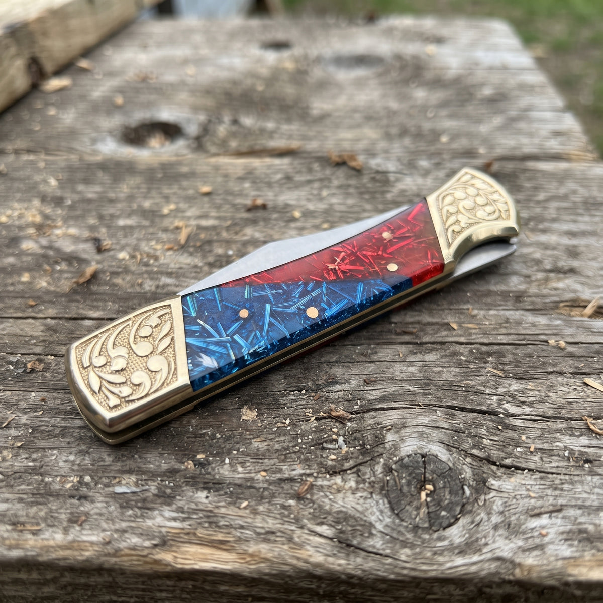 A folding pocket knife with a red and blue marbled inlay handle and ornate gold-toned metal end caps, resting on a weathered wooden surface.