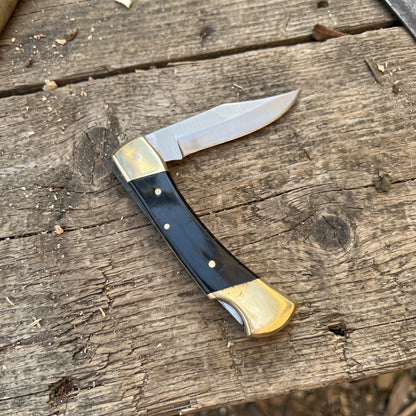 A folding pocket knife with a satin-finished steel blade and a black handle with brass bolsters and pins, resting on a rustic wooden surface.