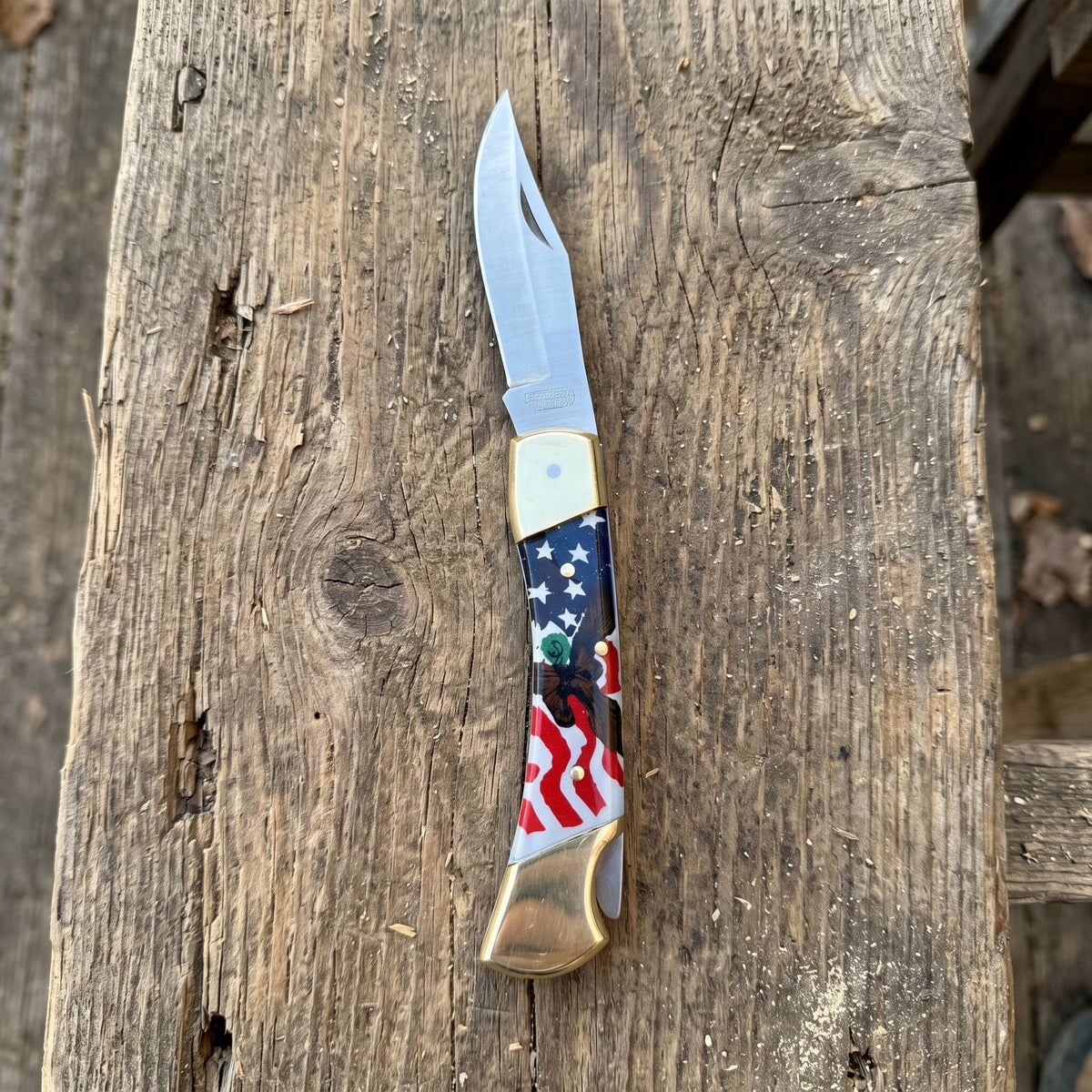 A folding pocket knife with a satin finish steel blade and a decorative handle featuring an American flag design, resting on a rustic wooden surface.