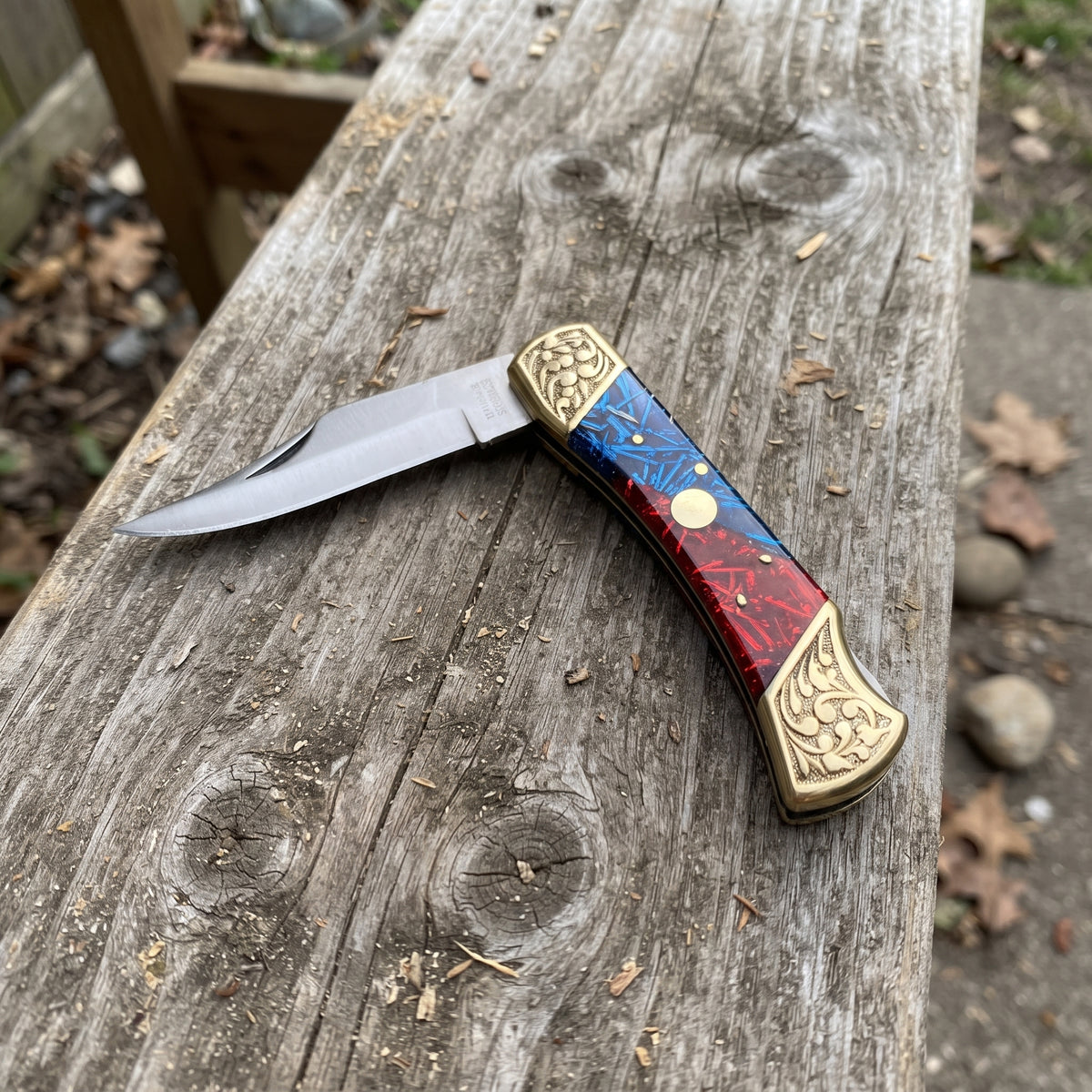 A folding pocket knife with a silver blade and a decorative handle featuring red, blue, and gold inlay, resting on a weathered wooden board outdoors.