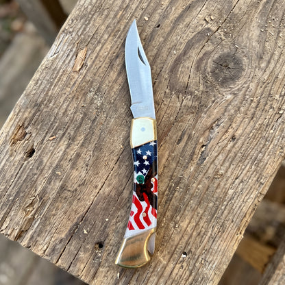 A folding pocket knife with a silver blade and a decorative handle featuring an American flag pattern, resting on a rustic wooden surface.