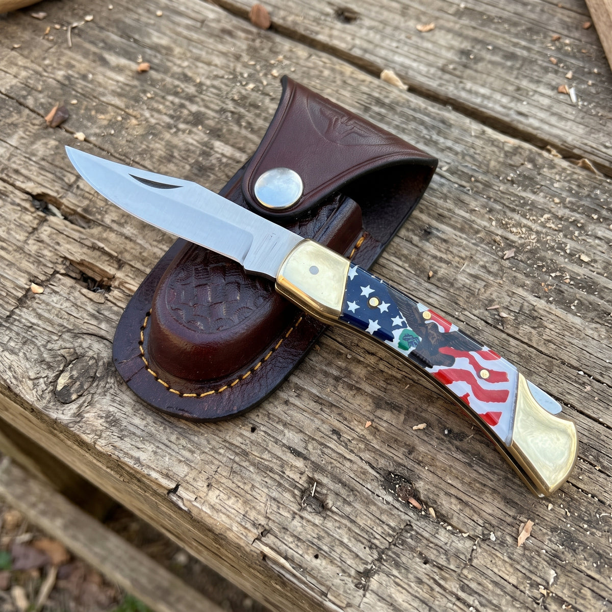 A folding pocket knife with a silver blade and a handle featuring an American flag pattern, resting on a brown leather sheath over a rustic wooden surface.