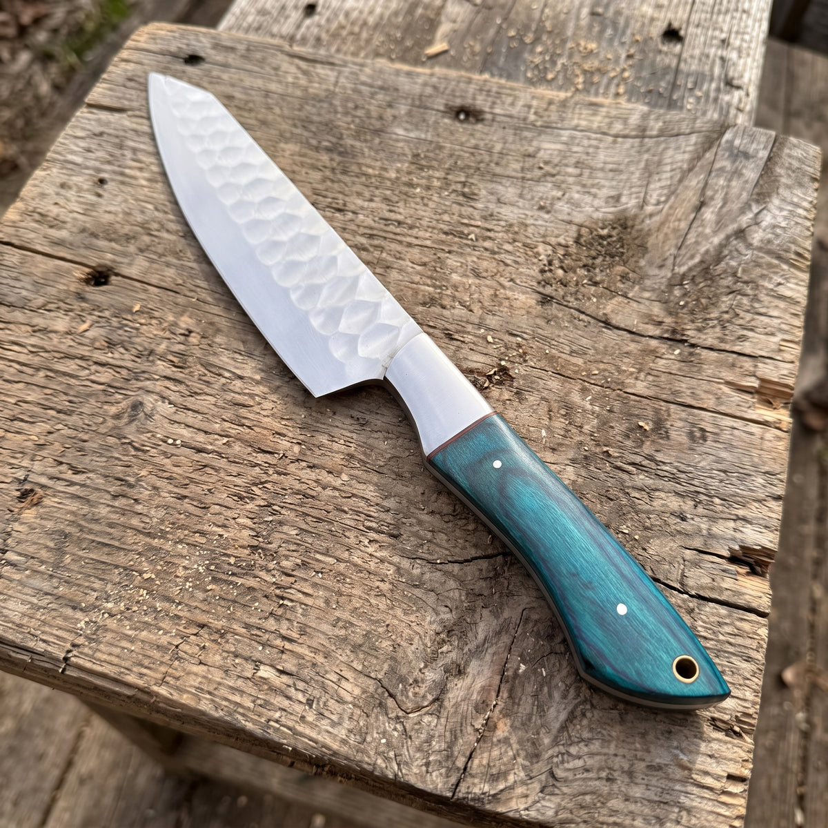 A kitchen knife with a hammered Damascus steel blade and a textured teal-green wooden handle, resting diagonally on a rustic weathered wooden plank.