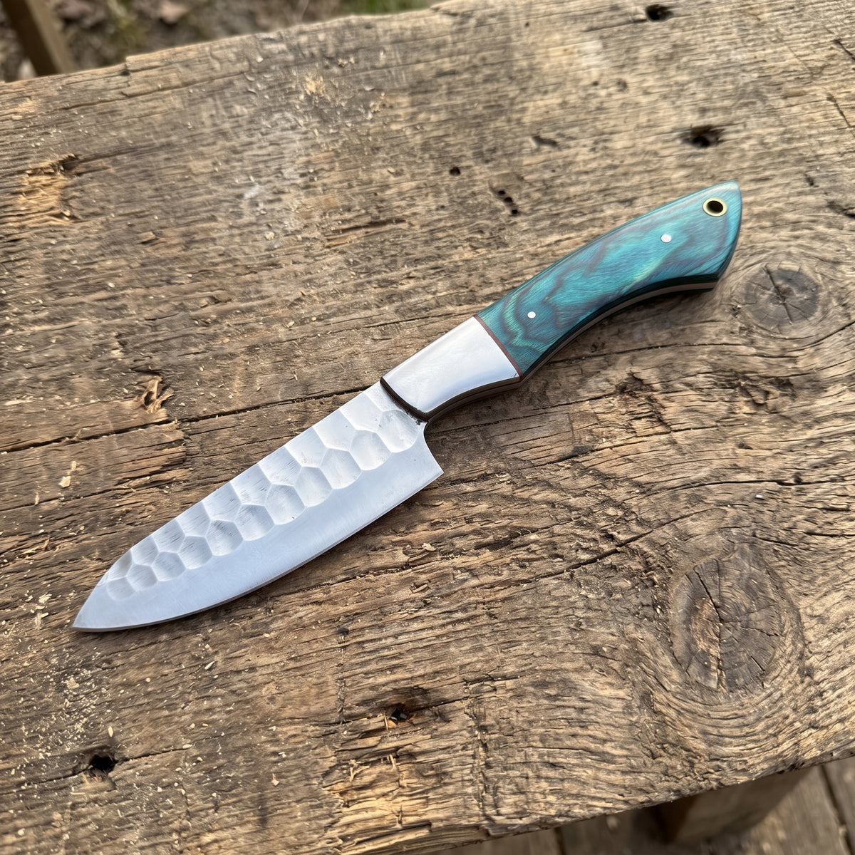 A fixed-blade chef knife with a hammered silver blade and a marbled turquoise-green handle, resting on a rustic weathered wooden surface.