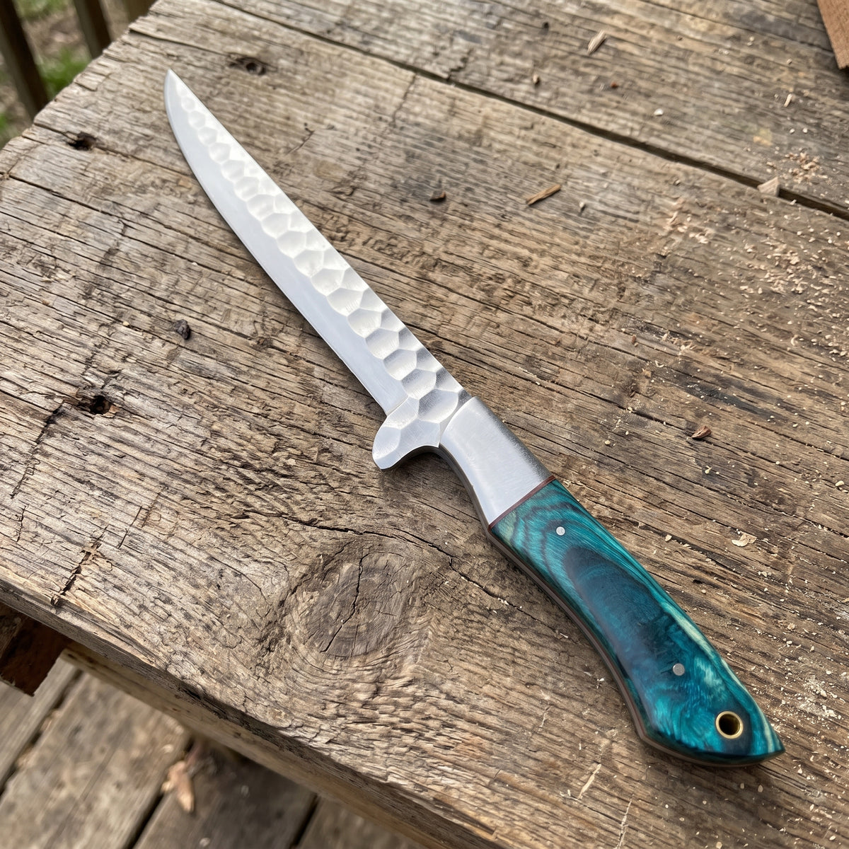 A fixed-blade chef knife with a hammered silver blade and a textured teal-green handle, resting diagonally on a rustic wooden plank surface.
