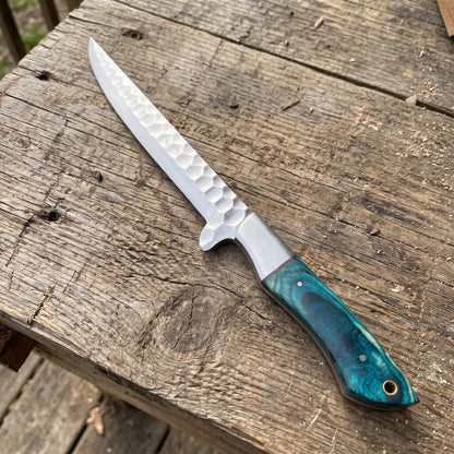 A fixed-blade chef knife with a hammered silver blade and a textured teal-green handle, resting diagonally on a rustic wooden plank surface.