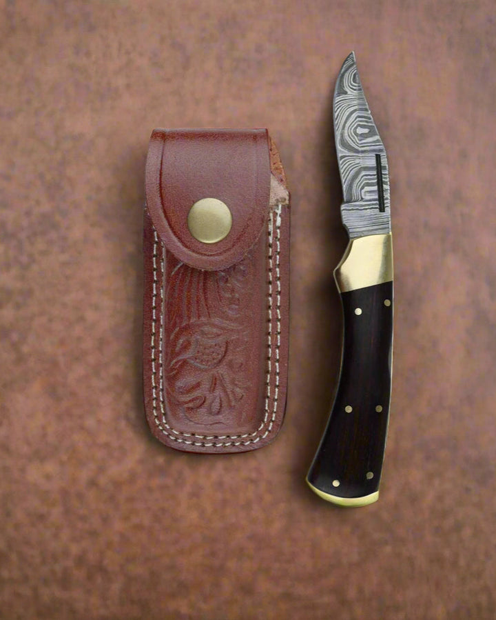 Hand-forged Damascus folding pocket knife with black wood handle and brass bolster beside a brown leather sheath on a textured surface