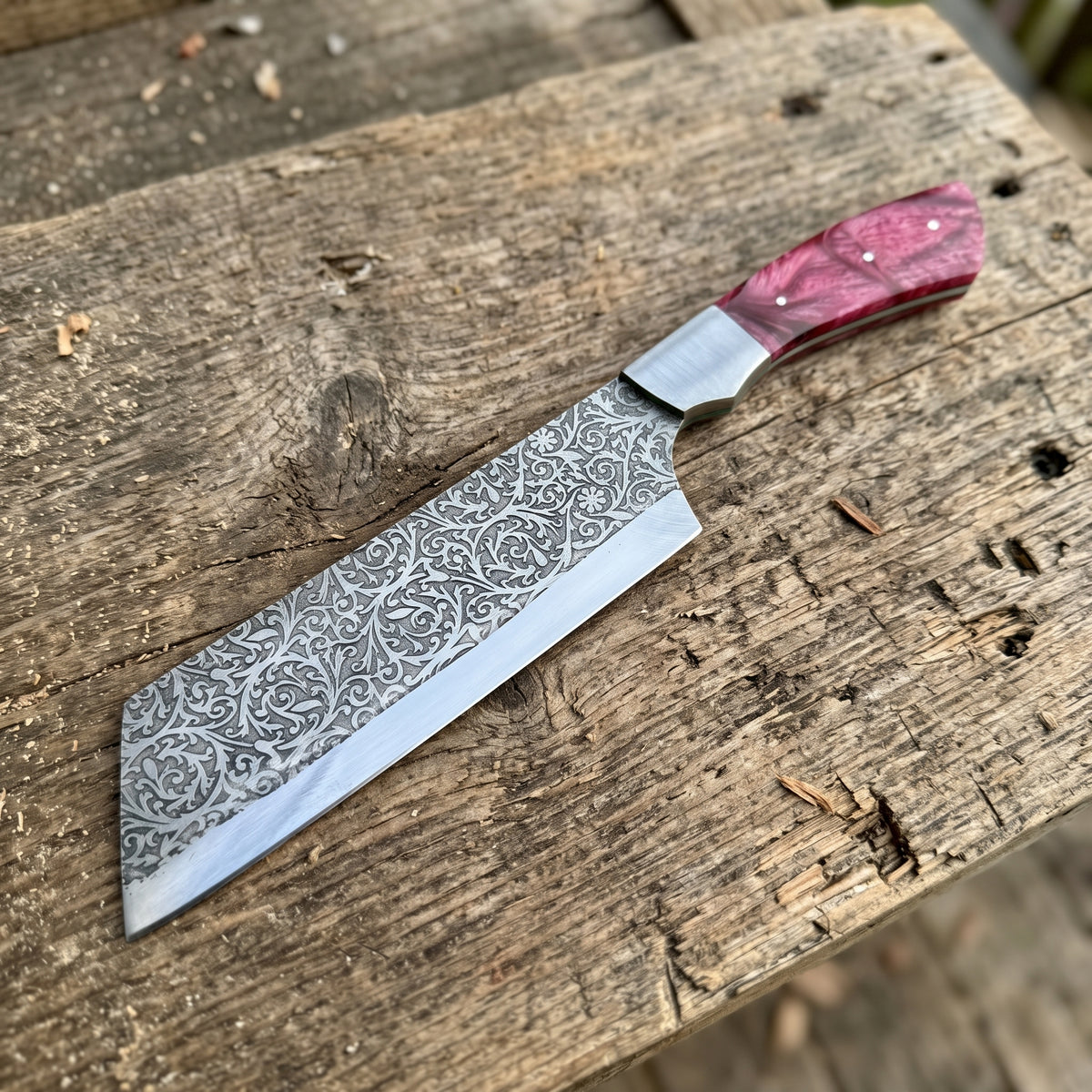 A handcrafted kitchen knife with a patterned Damascus steel blade and a pink marbled handle, resting on a rustic wooden surface.
