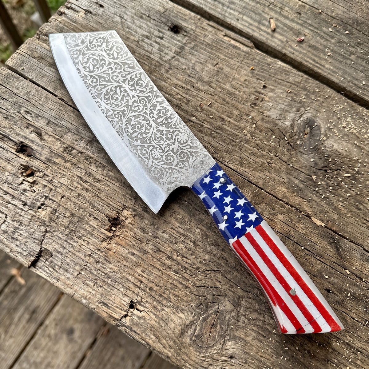 A kitchen knife with a patterned Damascus steel blade and a handle featuring an American flag design, resting on a rustic wooden surface.