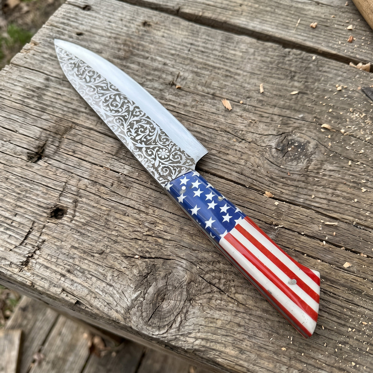 A kitchen knife with a patterned steel blade and a handle printed with the American flag, resting diagonally on a weathered wooden plank.