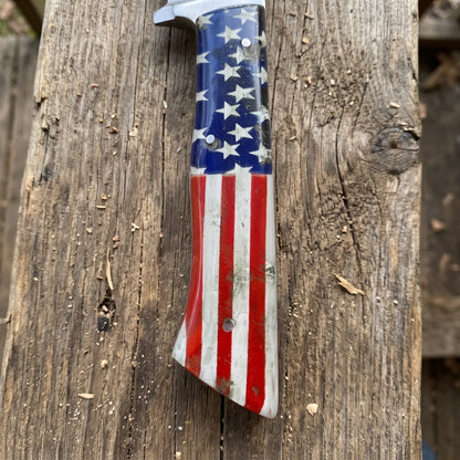 A knife handle featuring a painted American flag design with white stars on a blue background and red and white stripes, resting on a weathered wooden surface.