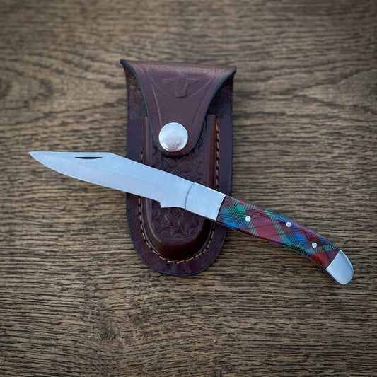 A old southern stainless steel pocket knife with a silver blade and a colorful tartan-patterned handle, displayed diagonally next to its brown leather sheath on a wooden surface.