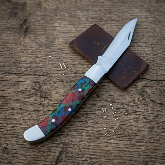 A old southern stainless steel pocket knife with a silver steel blade and a tartan-patterned handle in red, green, blue, and black, resting on a brown leather sheath over a rustic wooden surface.